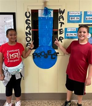 Students stand on either side of a poster of a water gauge with the words Ugandan Water Project on it